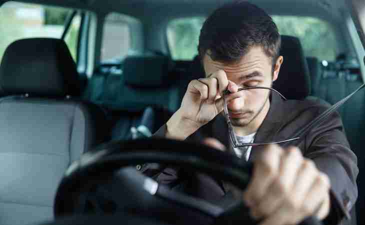 sleepy person rubbing eyes in car with hand on steering wheel